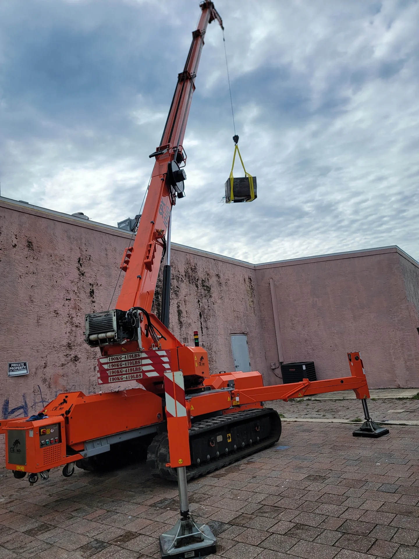 Orange tracked crane with deployed outriggers lifting a suspended unit beside a building.