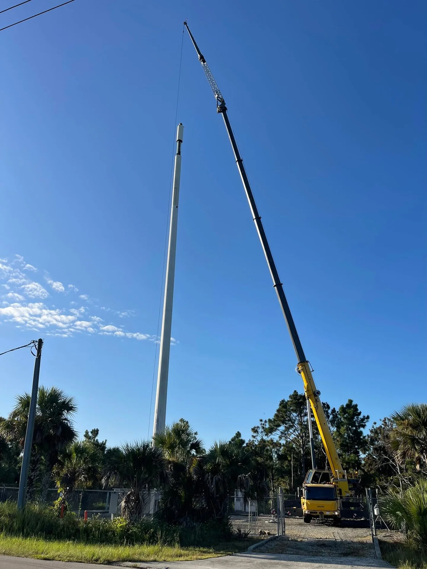Yellow crane with its boom extended high beside a suspended pole under a blue sky.