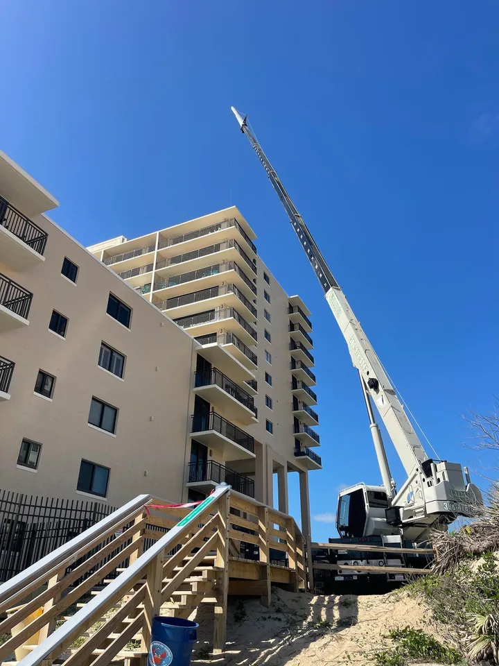 White crane with its boom raised beside a multi-storey building and wooden stairs.