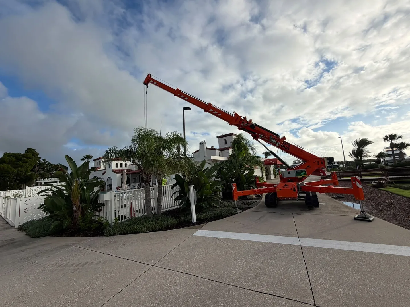 Orange compact crane set up beside white buildings and palms.