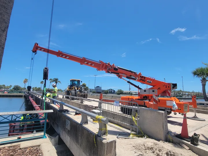Orange compact crane and people beside a waterfront work area.