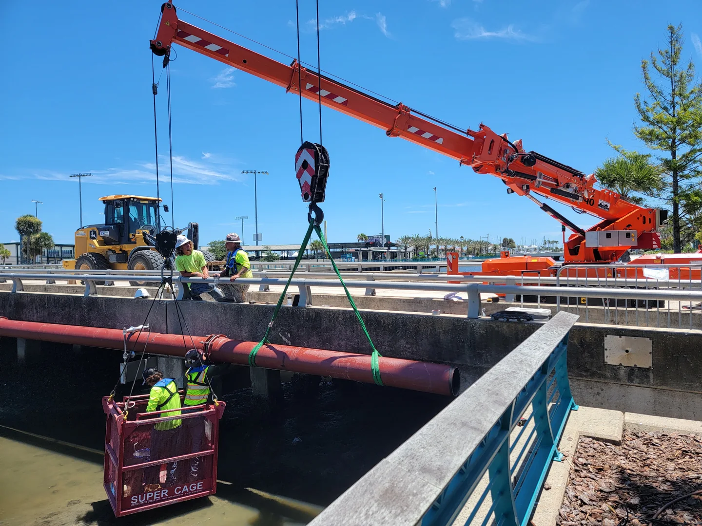 Crane hook and green slings attached to a large red pipe, with people beside the work area.