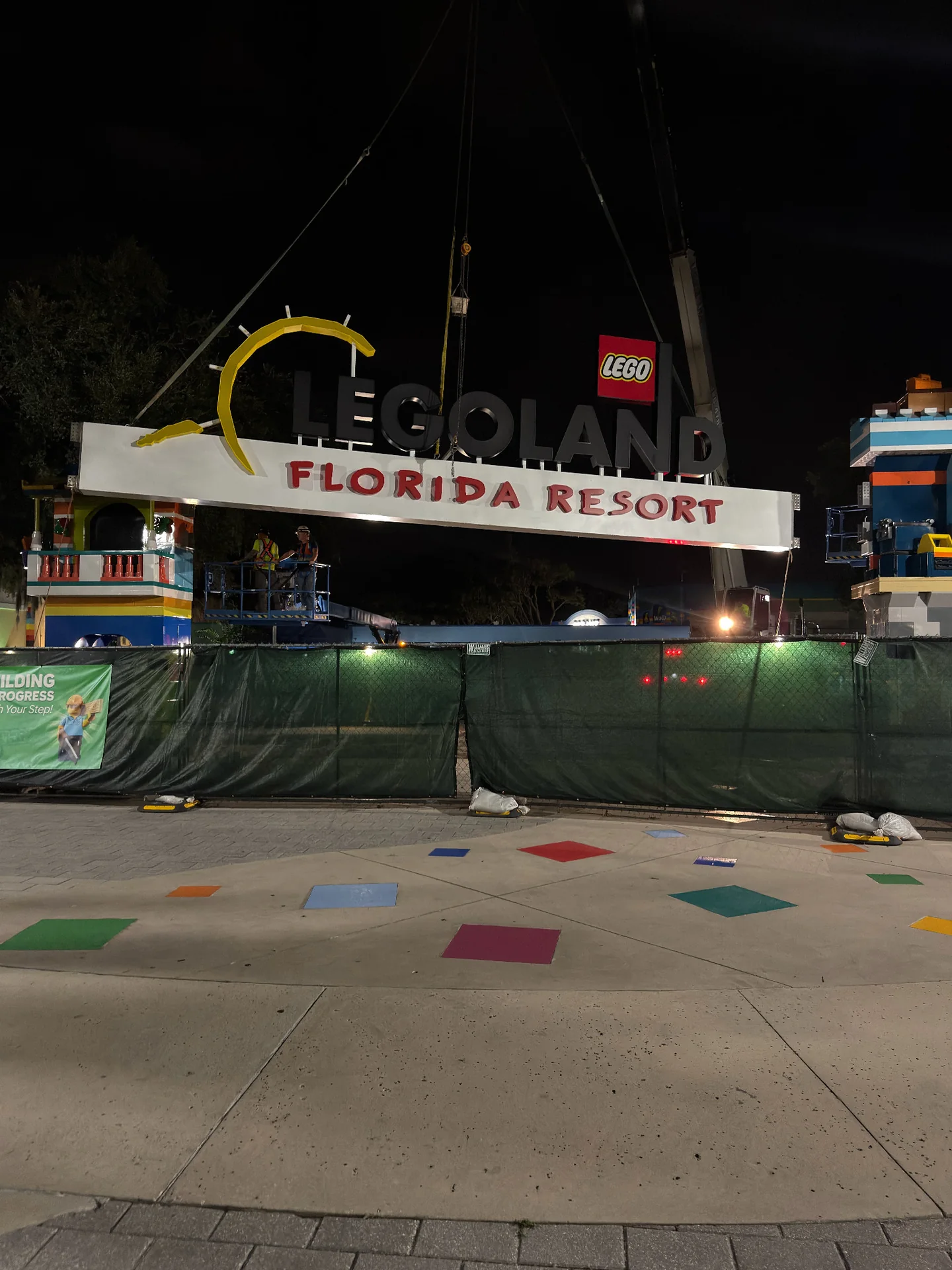 Crane rigging supporting a LEGOLAND Florida Resort sign at night, with people on an elevated platform.