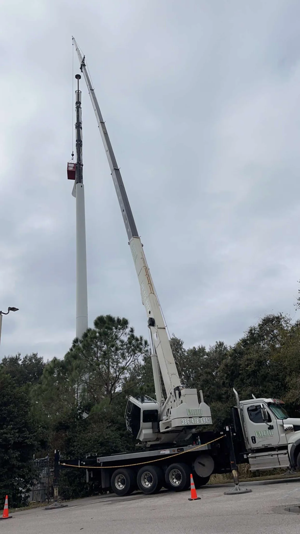 White crane truck with a raised boom beside a tall communications structure.
