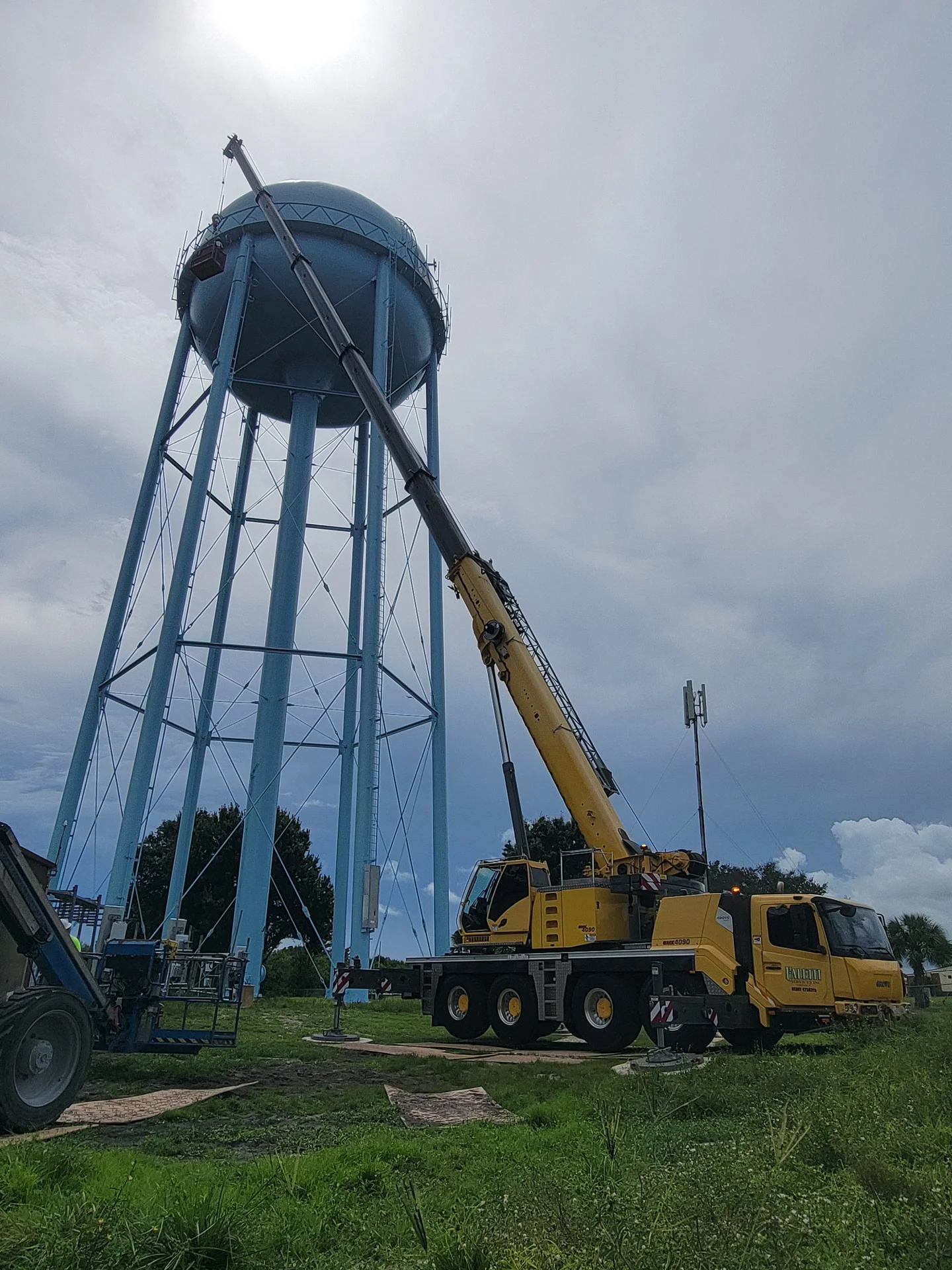 Yellow Faletti crane with its boom raised beside a blue water tower.